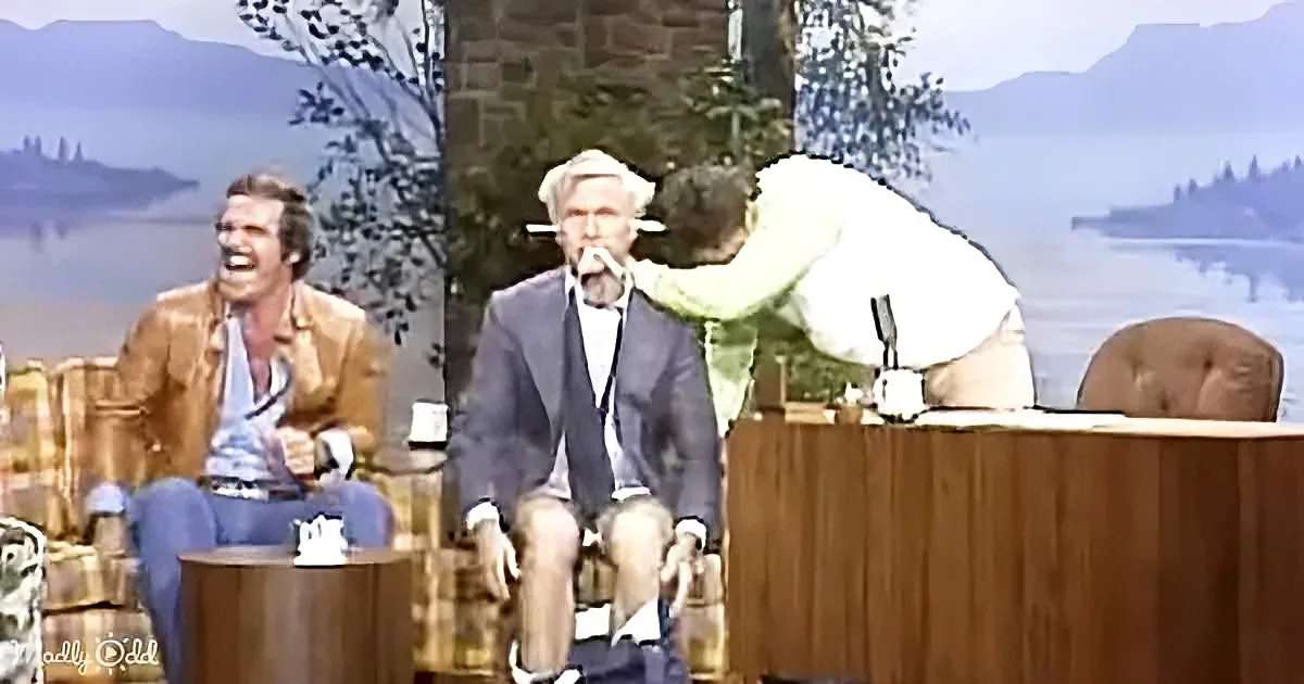A man in a suit is being tickled by another man on a talk show set with scenic background.