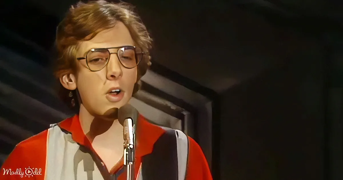 A person wearing glasses and a red shirt with white stripes stands in front of a microphone, singing.