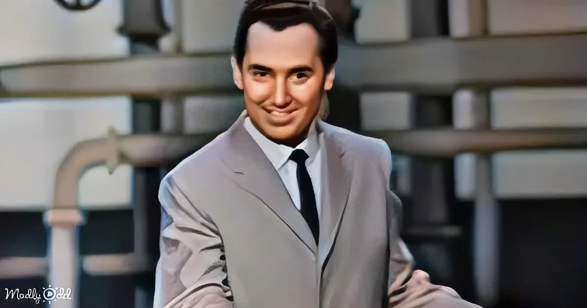 A man in a light gray suit and black tie smiles, standing in front of a blurred industrial background.