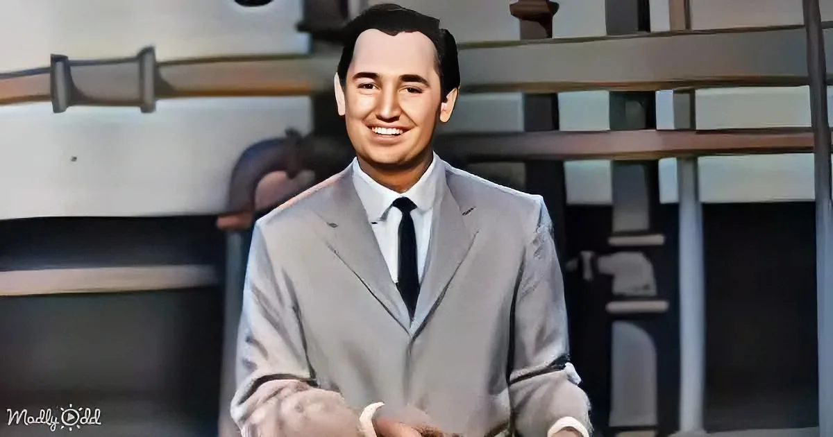 A man in a light gray suit and black tie smiles in a setting with a staircase and blurred background.
