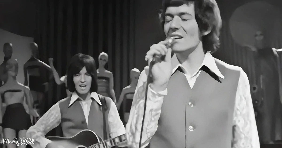 A man sings into a microphone, another plays guitar, both in retro attire, with mannequins in the background.