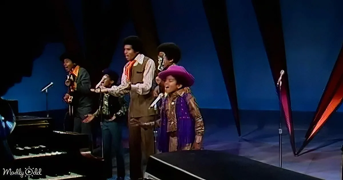 A young boy in a purple outfit sings on stage with a group, surrounded by microphones and a piano.