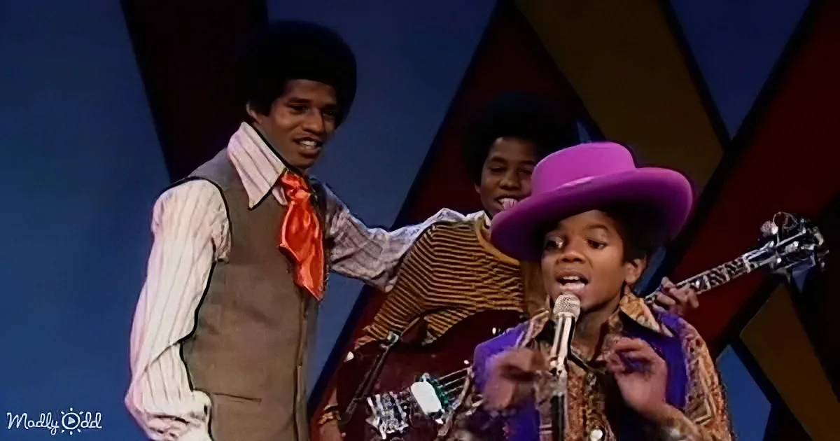 A young man in a purple hat sings into a microphone, accompanied by two other individuals on stage.