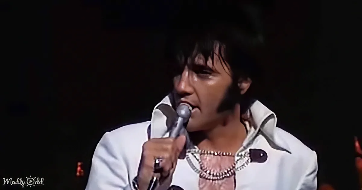 Elvis Presley in a white suit, holding a microphone, singing on stage.