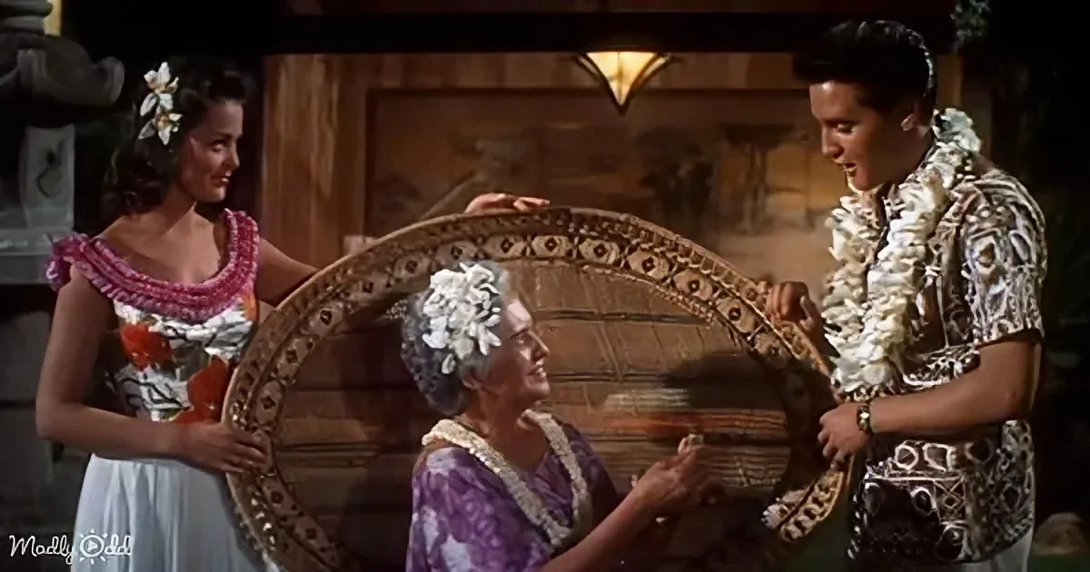 Elvis Presley and a woman in traditional attire hold a large, ornate fan, smiling at each other.