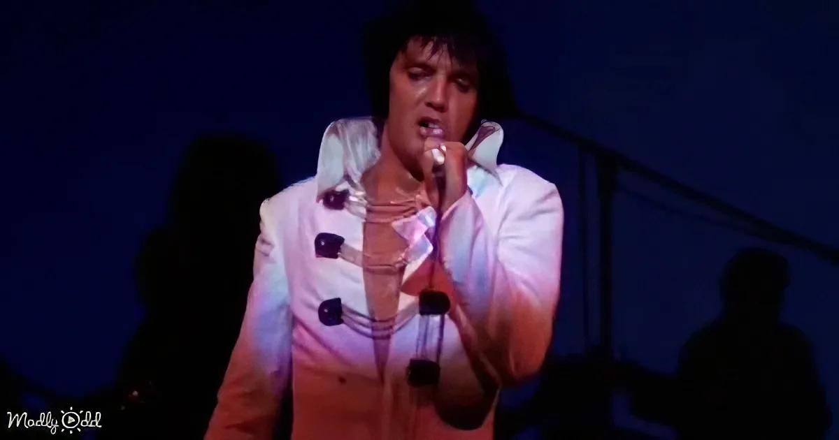 A performer in a white outfit sings into a microphone on stage under dim lighting.