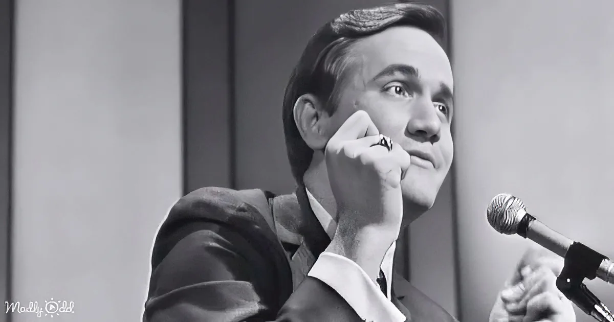 A man in a suit holds his hand to his face, looking up, with a microphone in front of him.