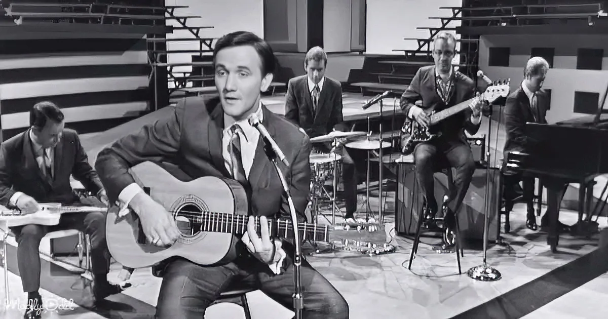 A man in a suit plays guitar on stage, singing into a microphone, with a band behind him.