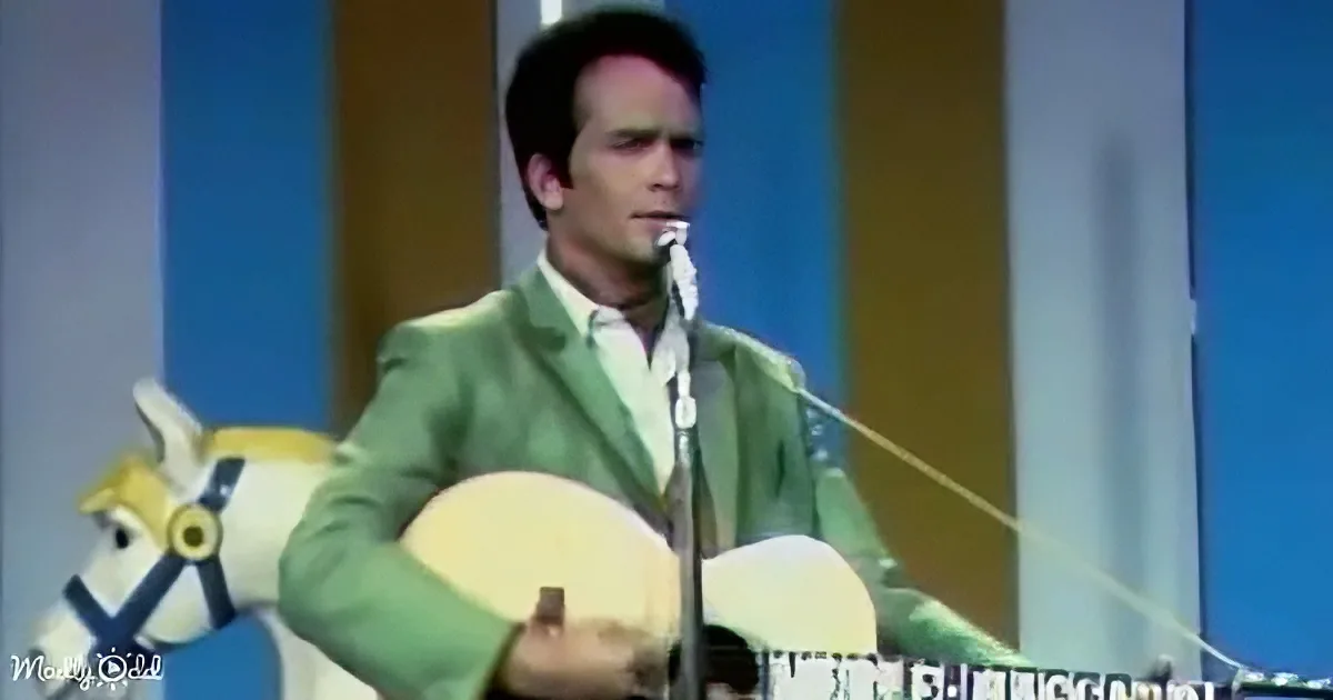 A man in a green suit plays a guitar on stage, with a microphone and a colorful background.
