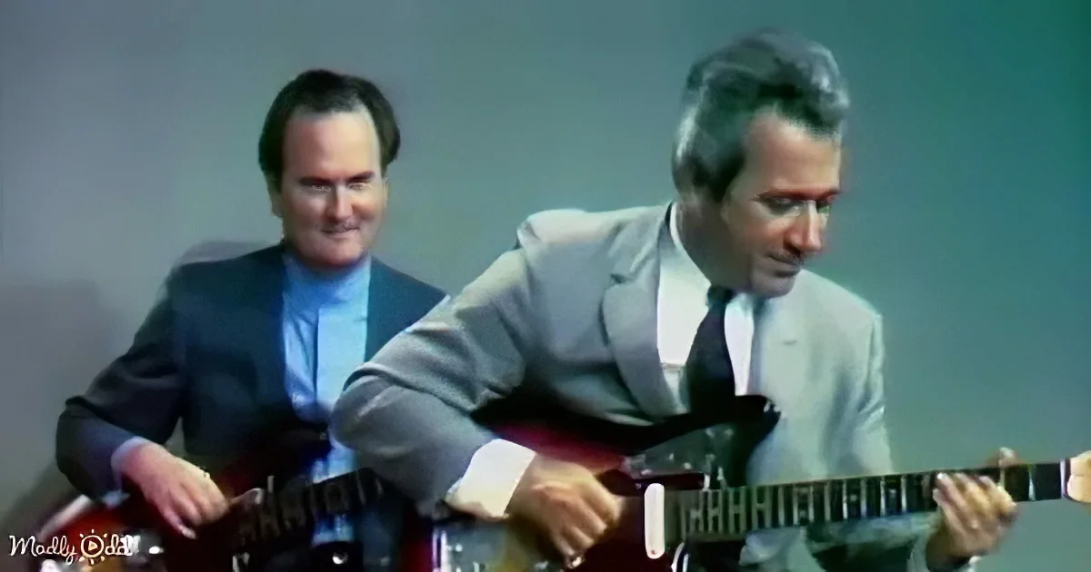 A man in a light suit plays a guitar, with another person in a dark suit playing a similar instrument behind him.