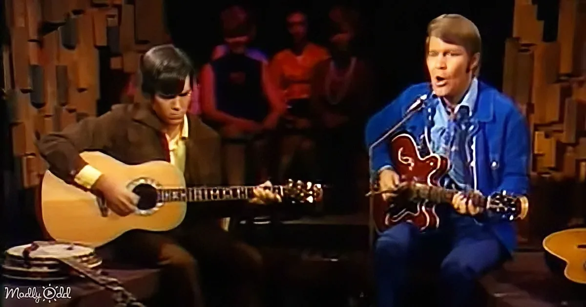 Two musicians play guitars on stage, one in blue, the other in brown, with an audience in the background.