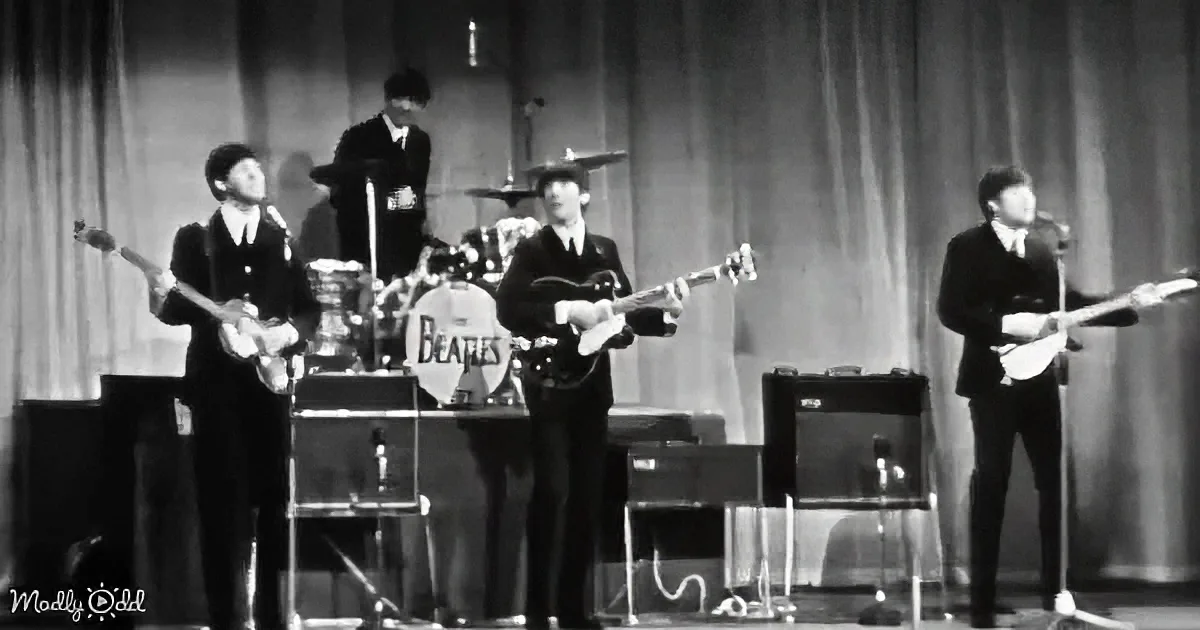 The Beatles perform on stage, dressed in suits, with a drummer and three guitarists, in a black-and-white image.