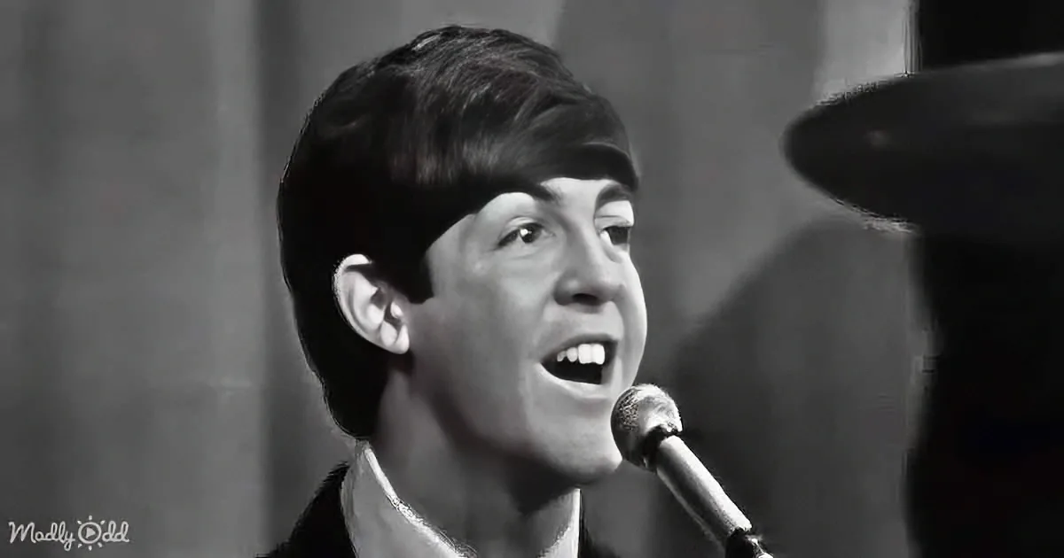 A man with short dark hair sings into a microphone, wearing a dark jacket and white shirt.