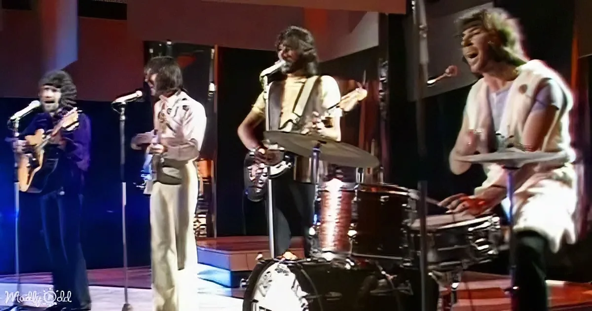 A band performs on stage with a drummer, guitarist, and singer, all wearing white and beige outfits.