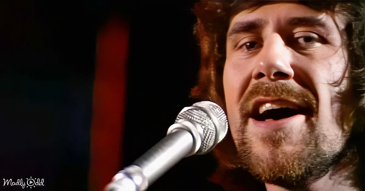 A man with curly hair and a beard sings into a microphone, his mouth open.