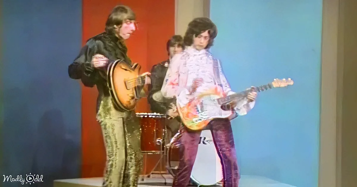 A musician in a colorful outfit plays guitar on stage with another musician in a black outfit.