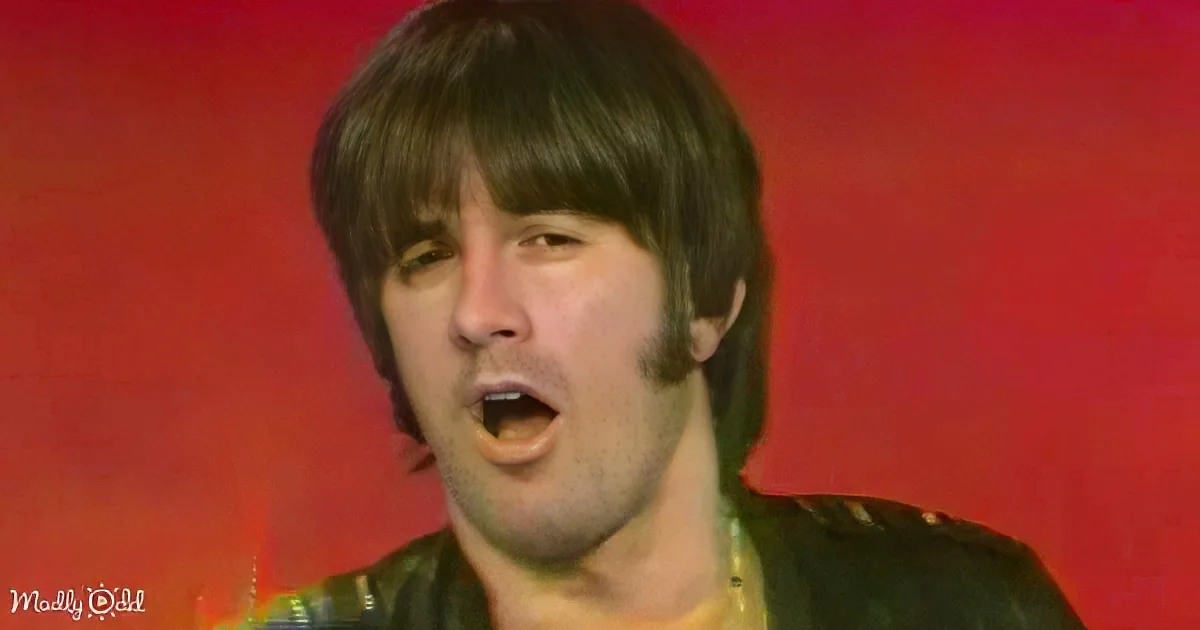A person with dark hair and a black leather jacket is singing against a red background.