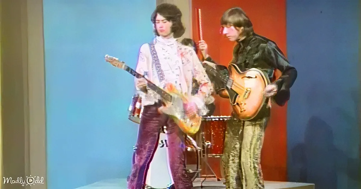 Two musicians play guitars on stage, one in a white outfit, the other in black, with a drum set in the background.
