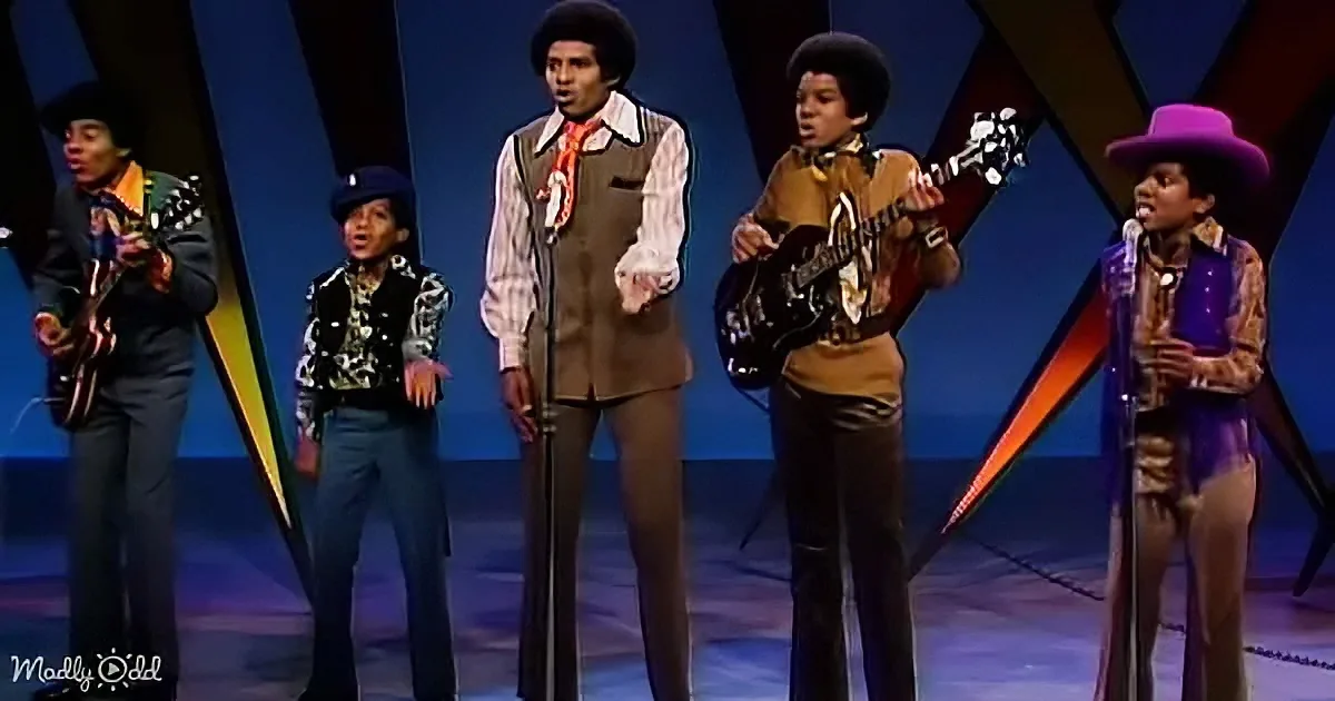 Five young musicians in 70s attire perform on stage with guitars and microphones.