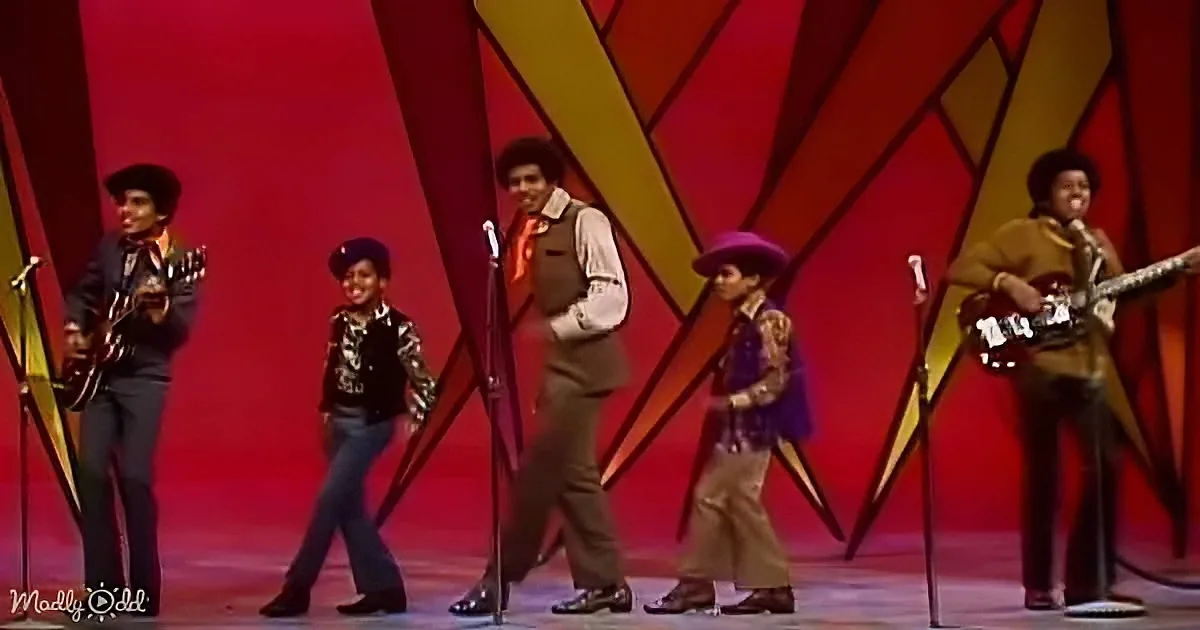 A group of musicians in vibrant outfits performs on stage with a red and yellow geometric backdrop.