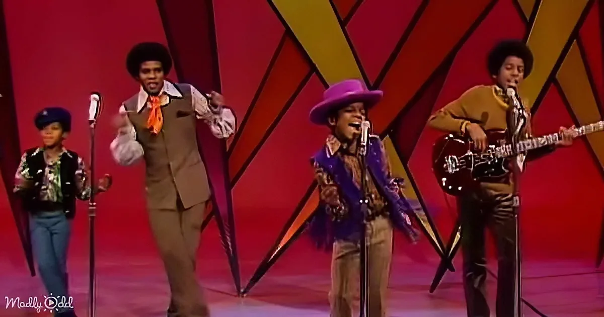 A group of performers in vibrant costumes sing and play instruments on a stage with a colorful geometric background.