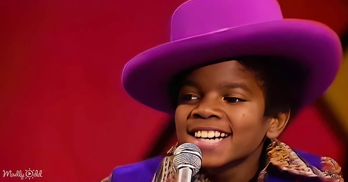 A smiling child wearing a purple hat and jacket, holding a microphone, with a red background.