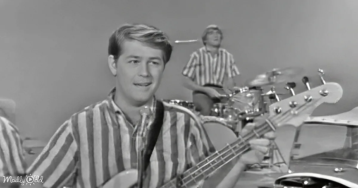 A man in a striped shirt plays a bass guitar, smiling, with a drummer in the background.
