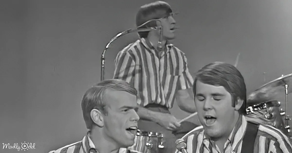 Two men in striped shirts sing passionately, accompanied by a drummer and a guitarist.