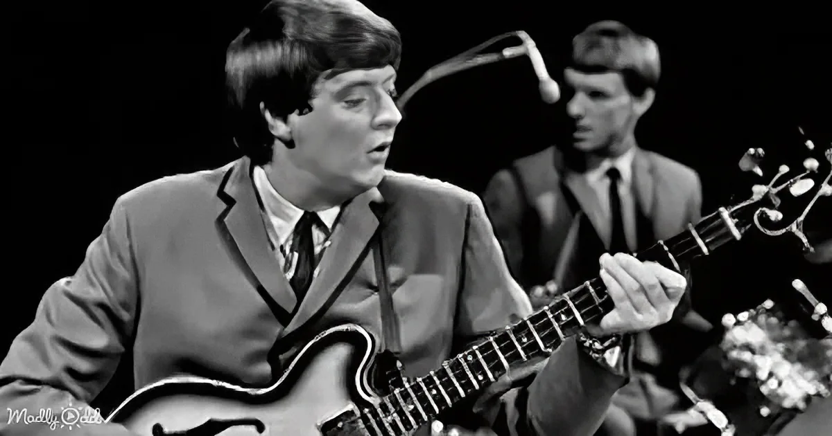 A man in a suit plays a guitar on stage, singing, with another musician in the background.