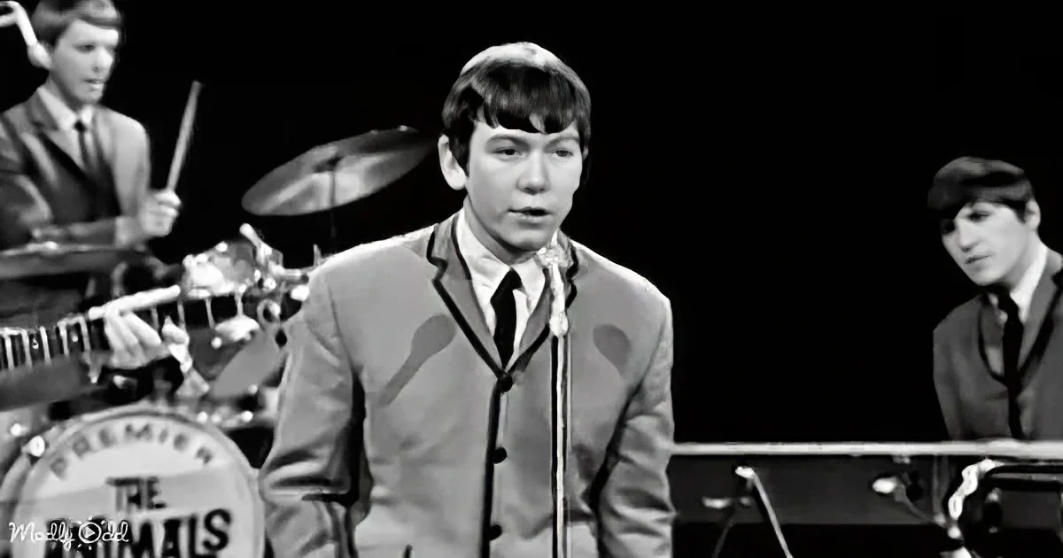 A young man in a suit sings into a microphone, with a drummer and keyboardist in the background.