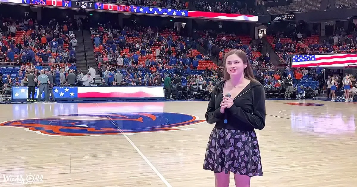A woman in a black dress stands on a basketball court holding a microphone, with a crowd and scoreboard in the background.