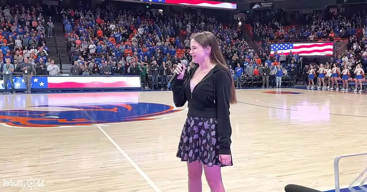 A woman in a black hoodie and skirt sings into a microphone on a basketball court with a cheering crowd and American flag...