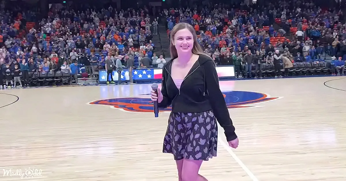 A woman in a black jacket and skirt holds a microphone on a basketball court with a crowd in the background.