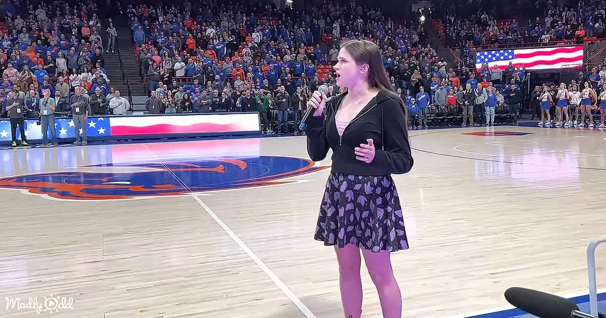 A woman sings on a basketball court with a large crowd and American flag in the background.