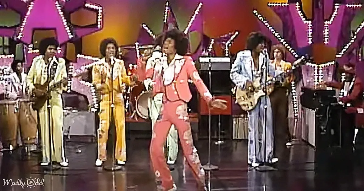 A man in a vibrant red suit dances energetically on stage with a band and colorful background.
