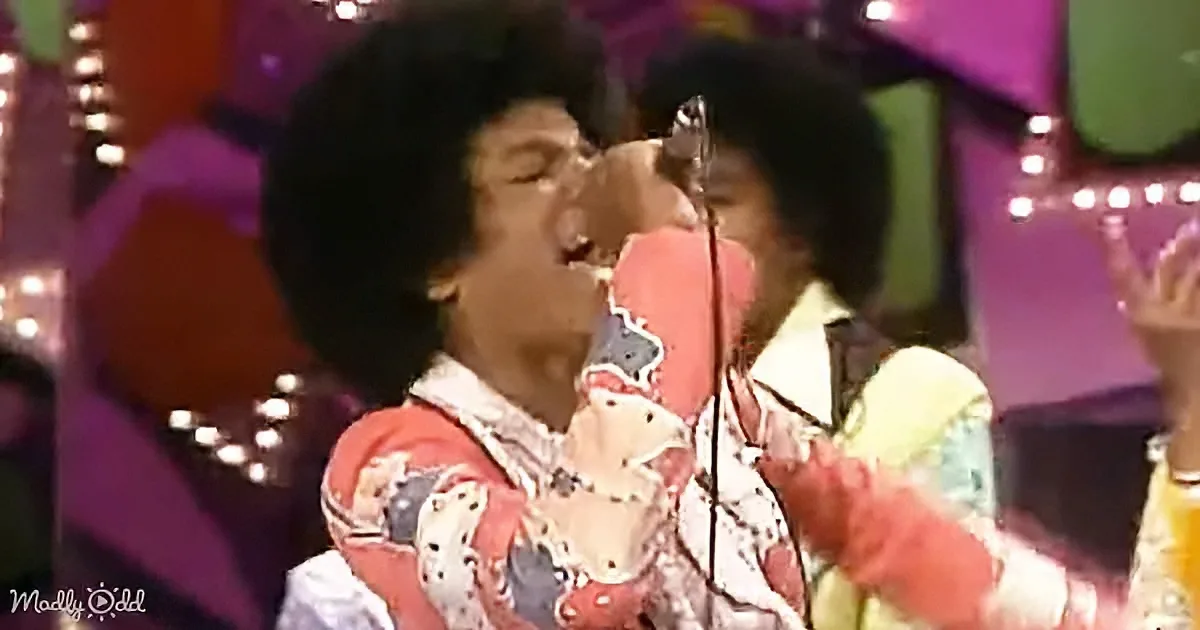 A person in a colorful outfit sings passionately into a microphone on a stage.