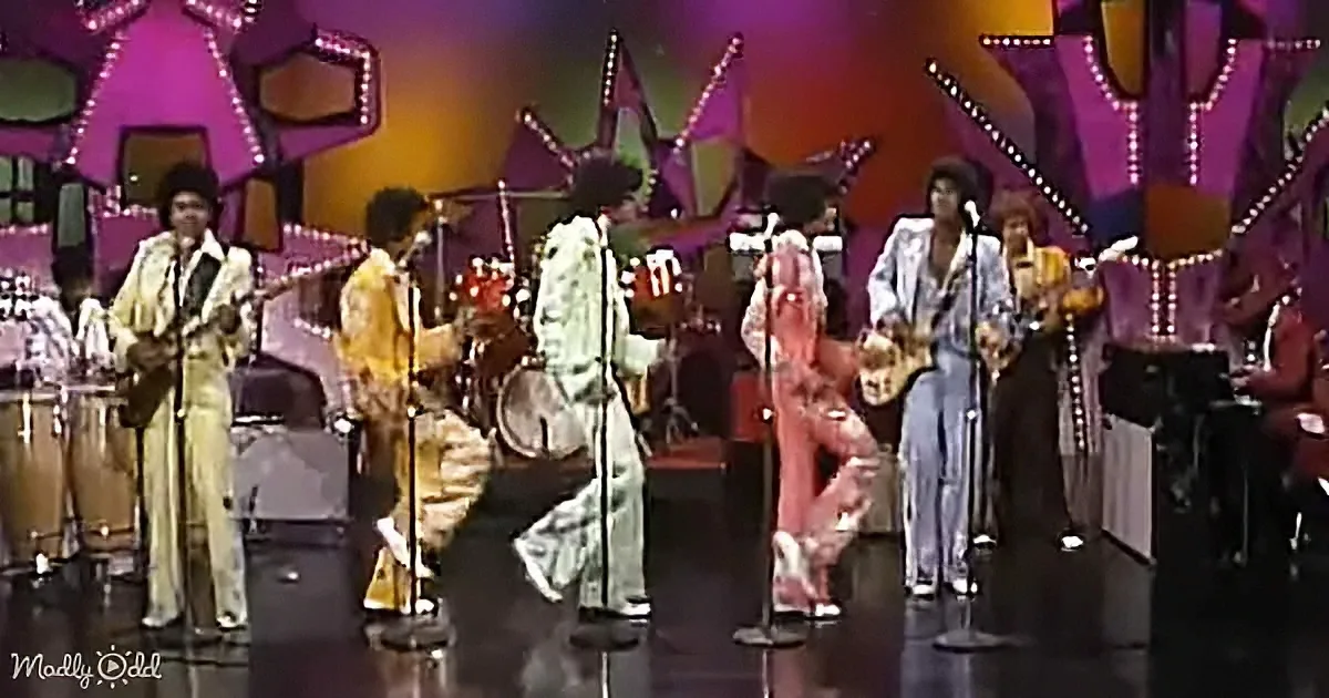 A vibrant stage performance featuring a group in colorful outfits, with a prominent figure in a pink outfit dancing...