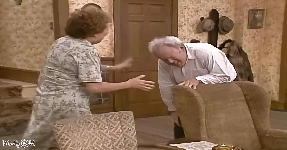 A woman in a floral dress is gesturing towards an older man leaning on a couch in a room with patterned wallpaper.