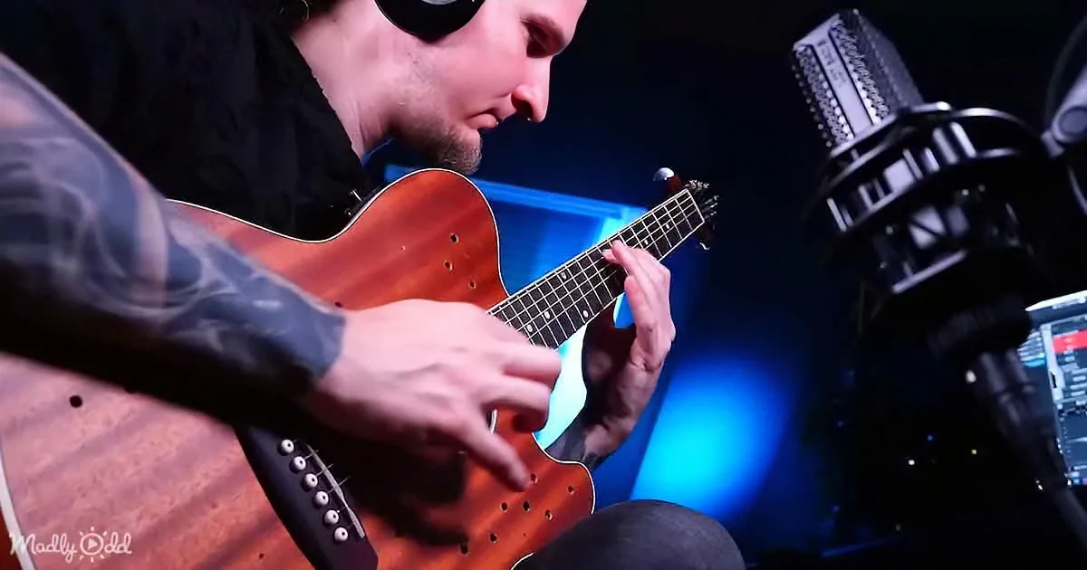 A man with tattoos plays an acoustic guitar in a dimly lit studio with a microphone and blue light.