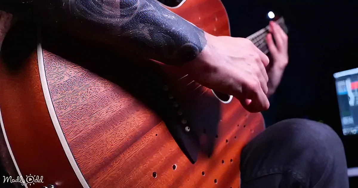 A tattooed arm strums a red acoustic guitar with a dark background.