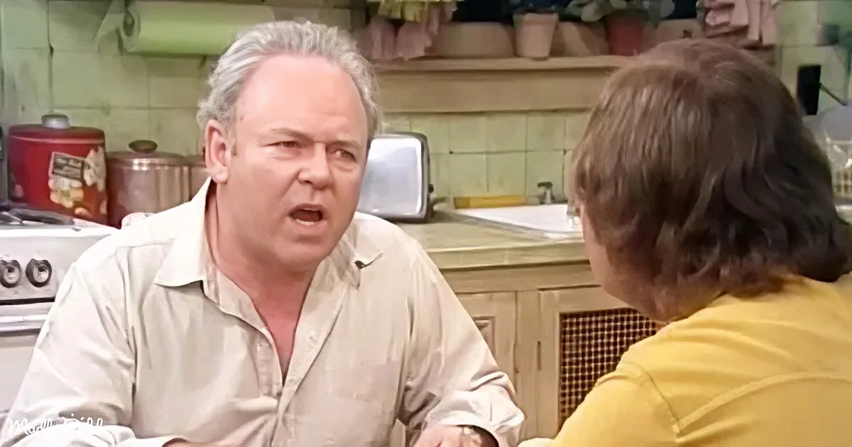 A man in a kitchen, wearing a light shirt, appears to be speaking or reacting to someone off-camera.