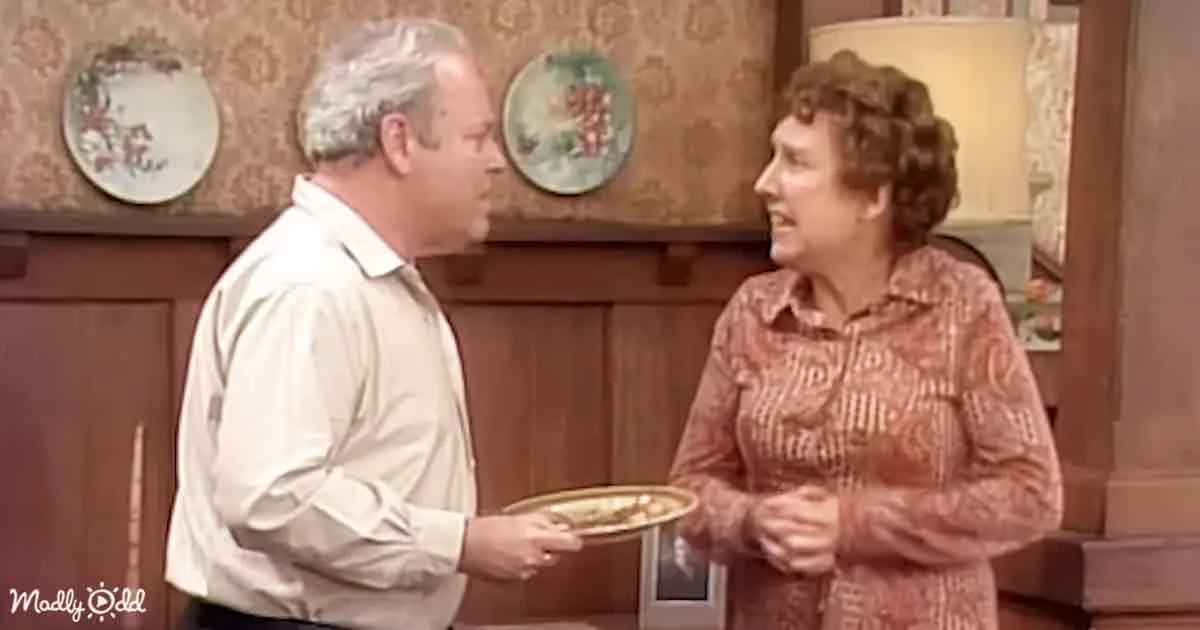 A man in a white shirt holds a plate, facing a woman in a patterned shirt in a kitchen setting.