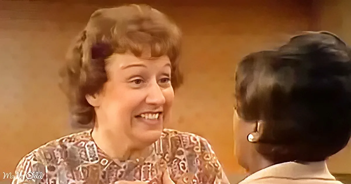 A smiling woman with curly hair and a patterned top is engaged in conversation with another person.