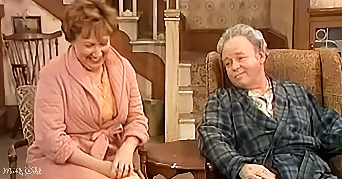 A woman in a pink dress sits beside a man in a plaid robe, both seated in a cozy, vintage-style room.