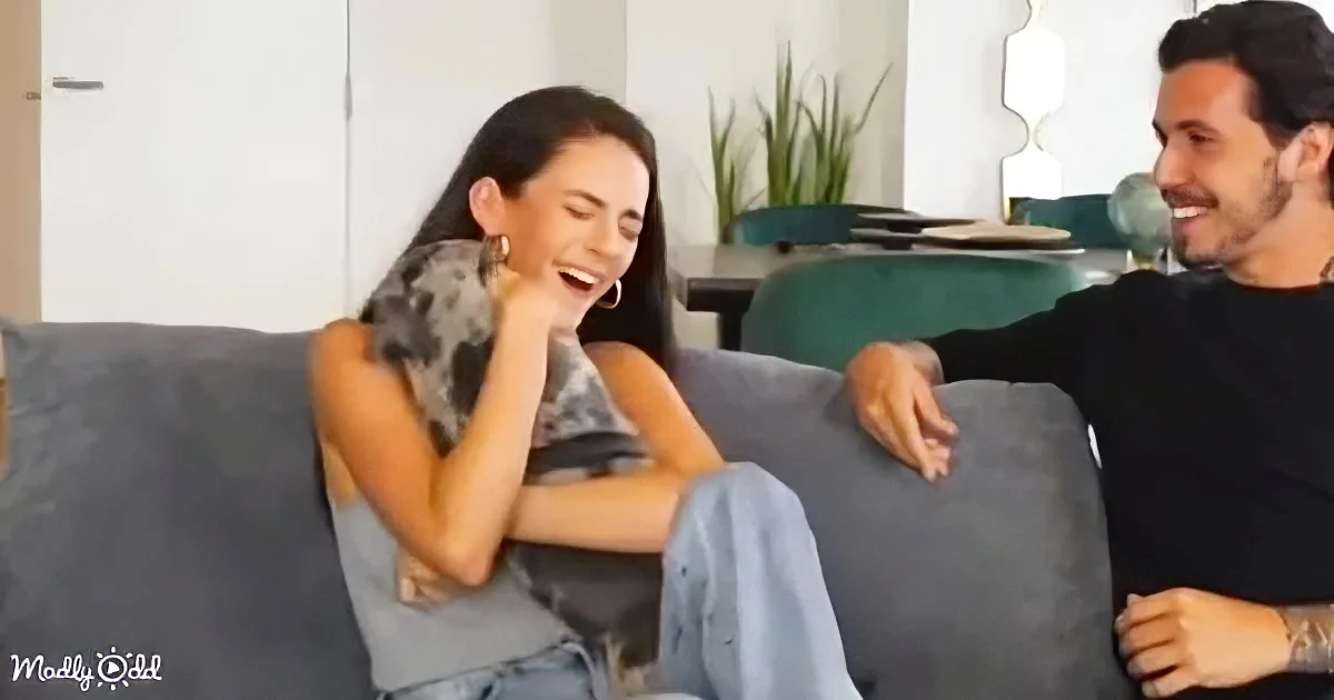 A woman laughs heartily on a couch, leaning on her hand, while a man sits beside her, smiling.