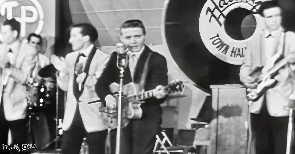 A man plays guitar on stage with a microphone, surrounded by other musicians and a Town Hall sign.