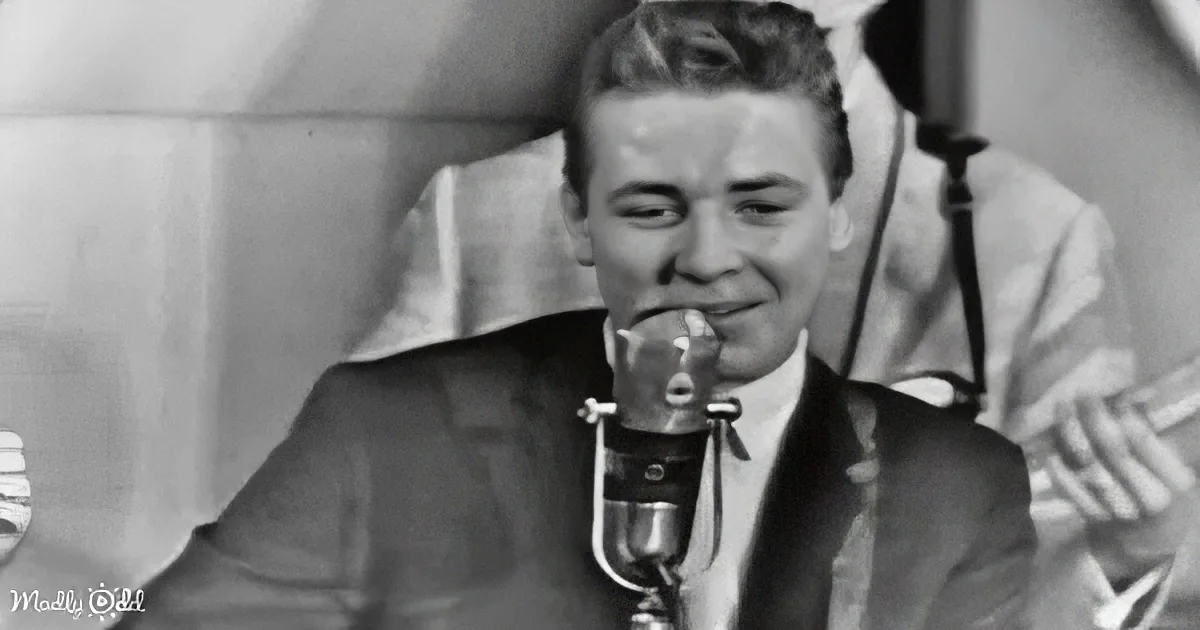 A man in a suit smiles into a microphone, seated with a musical instrument in the background.