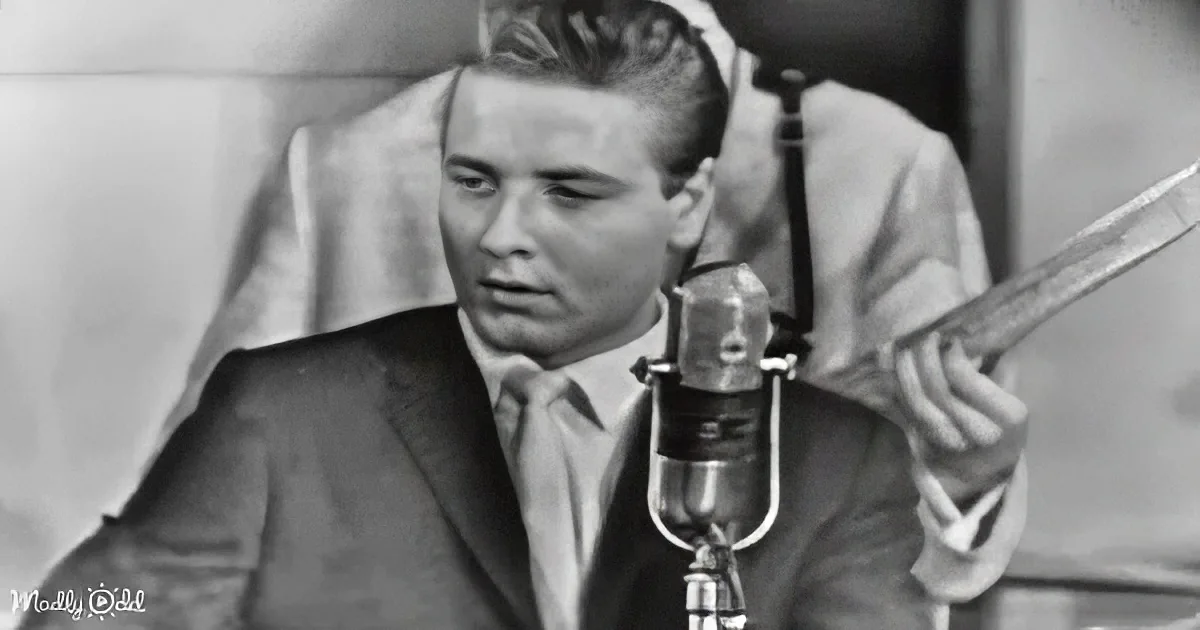 A man in a suit and tie sits at a microphone, holding a conductor's baton, suggesting a radio or recording session.