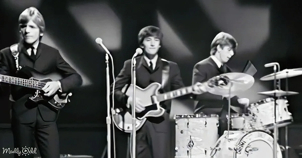 The Beatles perform on stage, dressed in suits, with microphones and instruments, under spotlight.