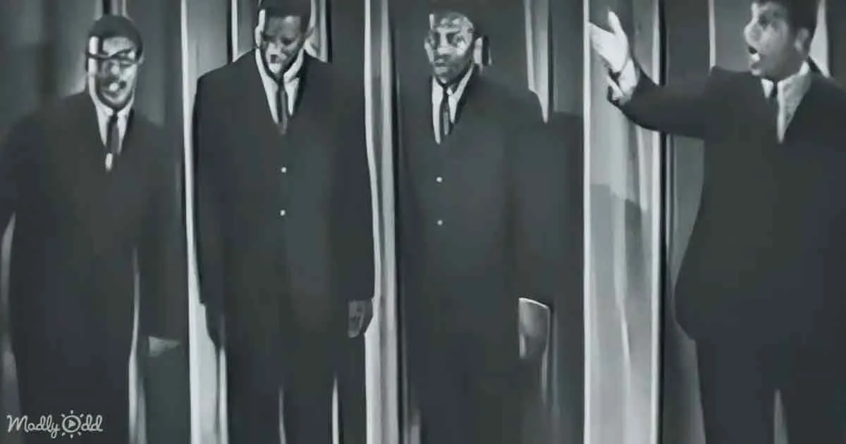 A man in a suit gestures dramatically while three others in suits stand in a line, all in black and white.